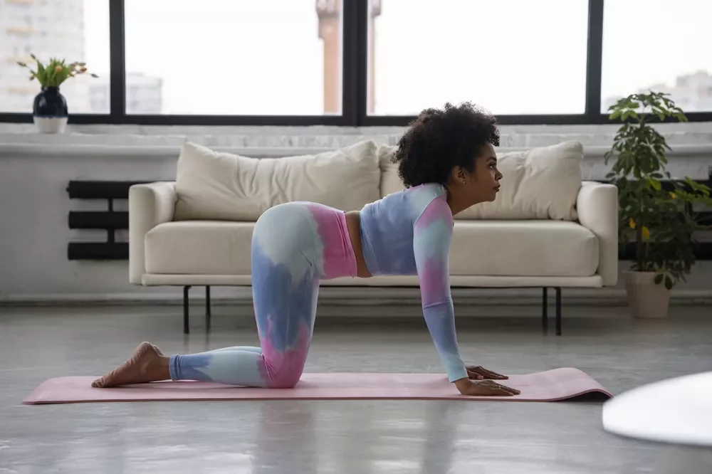 Vibration Plate Exercises For Sciatica: How To Use Them Safely For Real Relief 9 A woman with curly hair wearing tie-dye activewear performing a cat-camel spinal mobility stretch on a pink yoga mat in a living room.