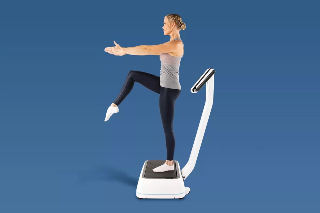 Top Vibration Plate Exercises: A Complete Guide To Safe Whole-Body Vibration Workouts 19 Woman practicing a single-leg balance pose on a vibration platform with arms extended forward for stability.