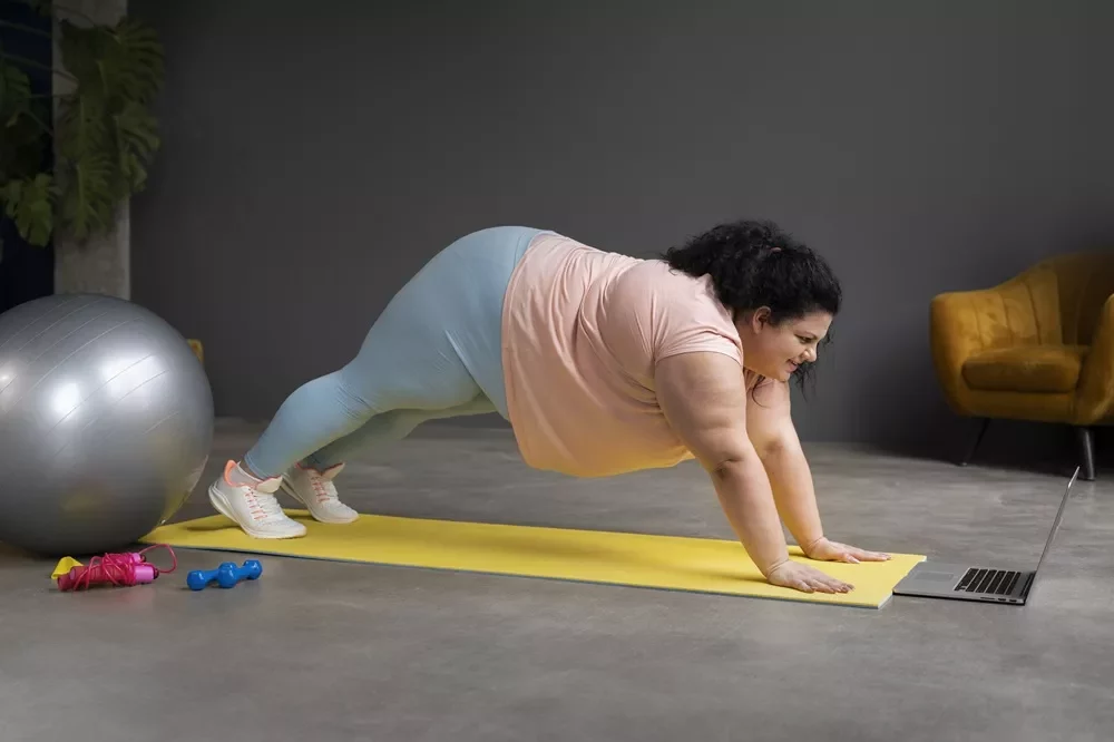 Best Vibration‑Plate Exercises for Belly Fat: Science‑Backed Tips, Workouts & Results 8 A woman in a pink t-shirt and light blue leggings performing a high plank on a yellow yoga mat while following a workout on a laptop.