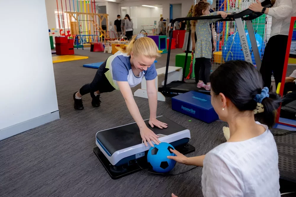 child vibration plate plank exercise A young girl performing a plank on a Hypervibe vibration plate while interacting with a therapist and a blue soccer ball in a clinical setting.