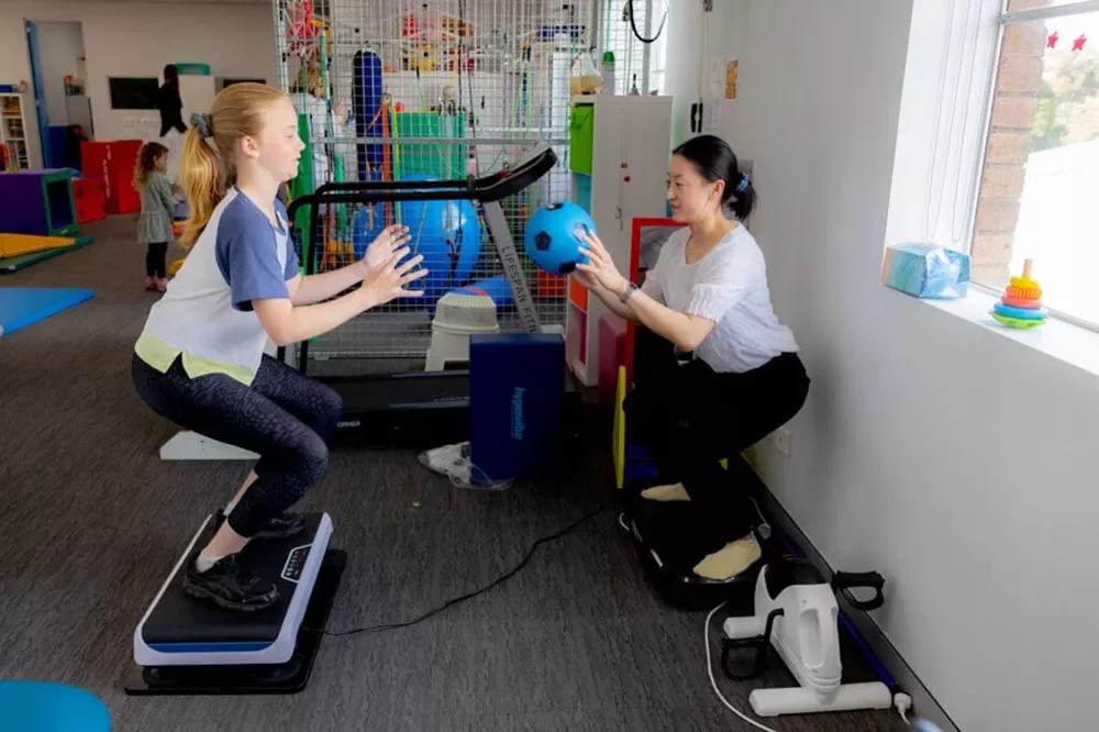 pediatric vibration squat coordination drill Pediatric therapy session showing a girl in a squat position on a vibration machine playing catch with a blue ball.