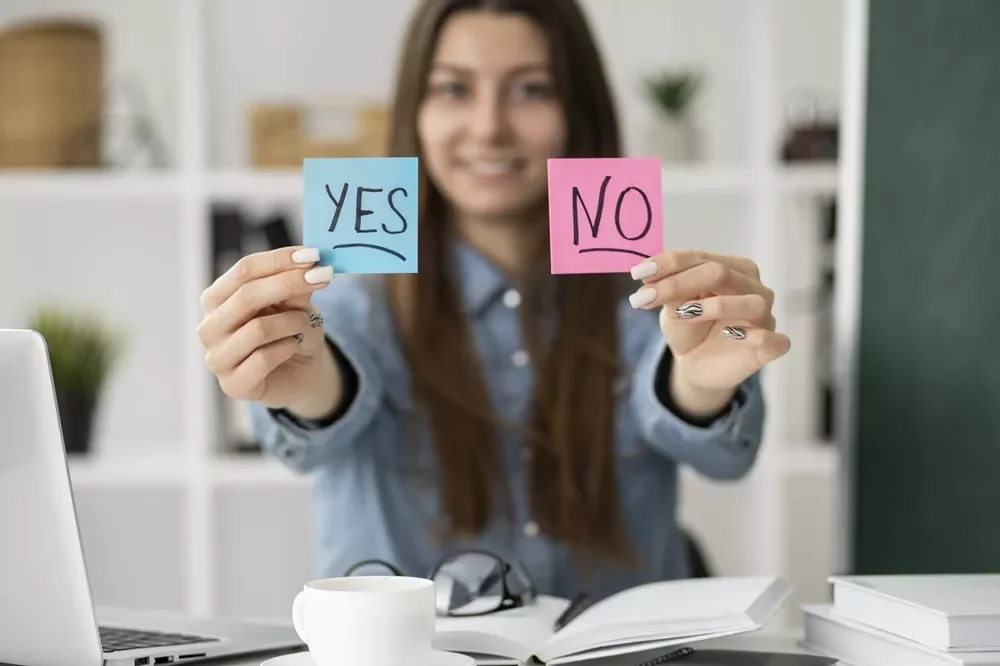 oman holding yes no sticky notes decision making Smiling woman at a desk holding blue and pink sticky notes that say YES and NO toward the camera.