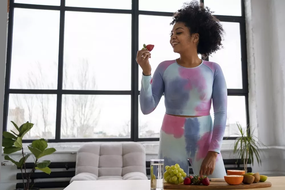 Woman in colorful activewear eating a strawberry while standing by a table of fresh fruit.