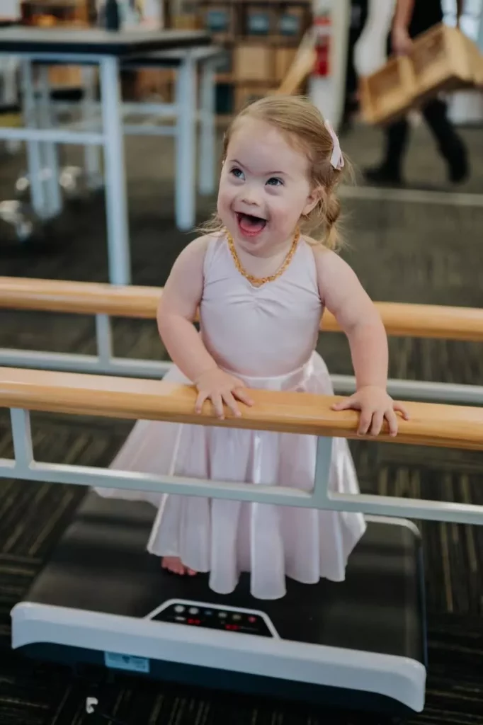 adaptive pediatric vibration barre training Happy young girl in a pink dress standing barefoot on a vibration machine utilizing a double ballet barre for balance