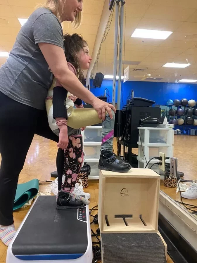 cerebral palsy vibration step up exercise Young girl with leg braces performing a supported step-up exercise on a vibration plate and wooden box