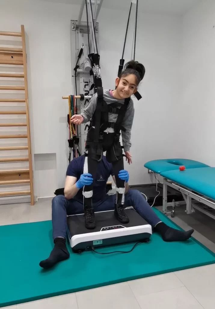 harness supported pediatric vibration therapy Smiling young girl supported by a overhead harness system while standing on a vibration machine in a clinic