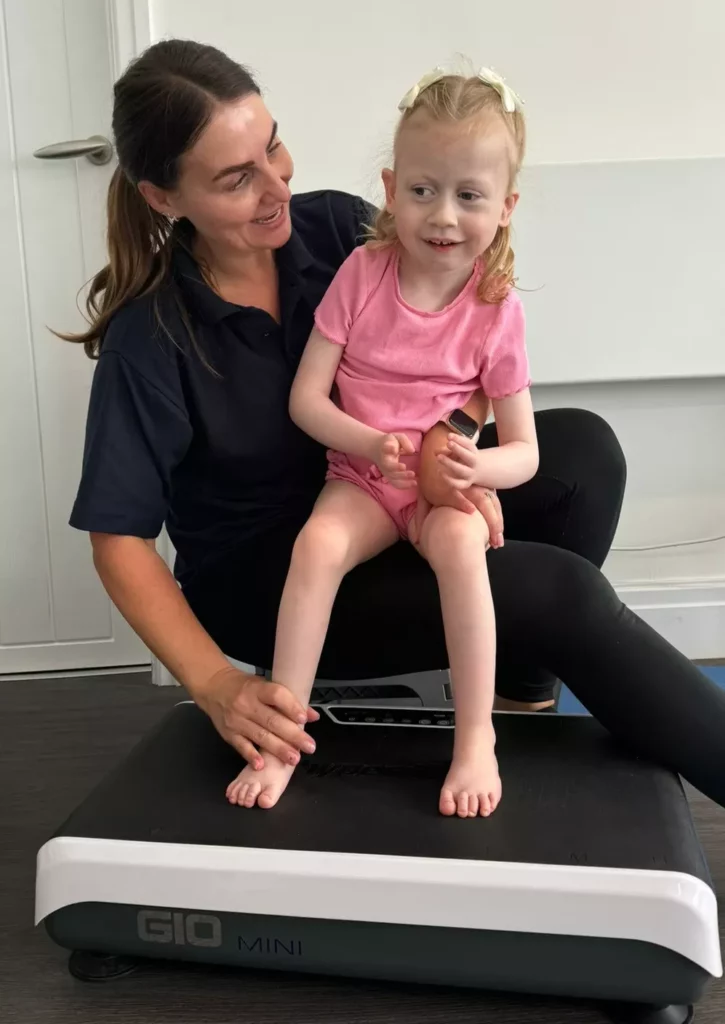 seated pediatric vibration foot stimulation Physical therapist supporting a young girl sitting on her lap with her bare feet placed on a Gio Mini vibration platform