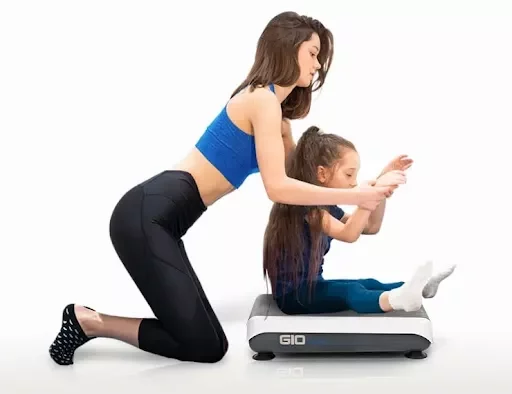 child sensory vibration plate therapy A young girl sitting on a Hypervibe G10 vibration plate while a female instructor assists with her posture.