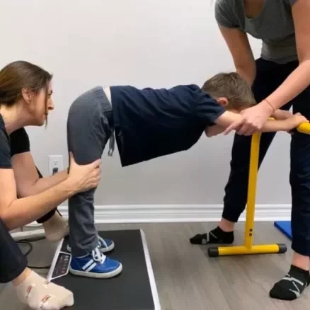 pediatric vibration plate exercise support A boy performing a leaning exercise on a vibration plate while two adults provide physical support and guidance.