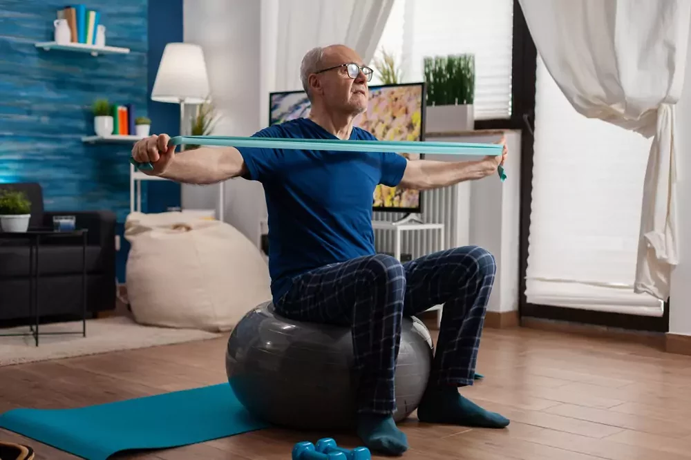 A senior man sitting on a ball, using a resistance band