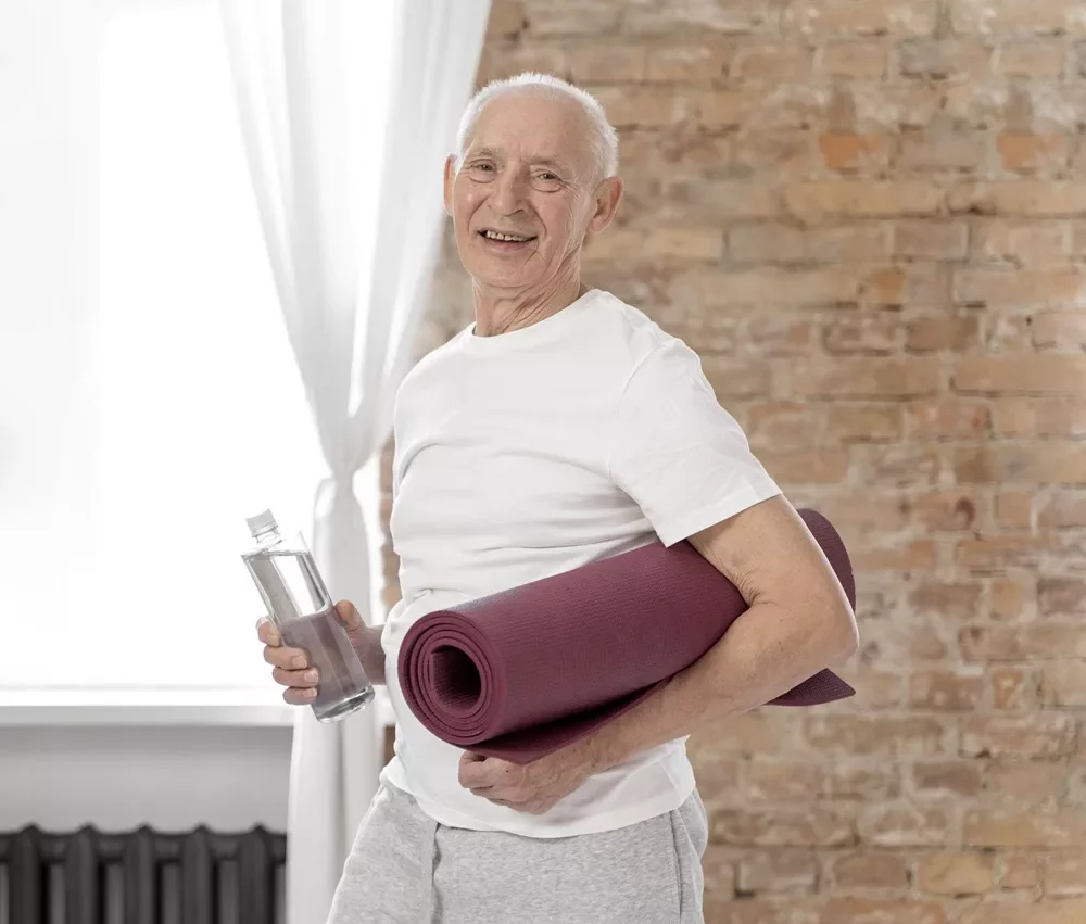 senior man with yoga mat posture fitness Smiling older man holding a rolled yoga mat and a water bottle in front of a brick wall.